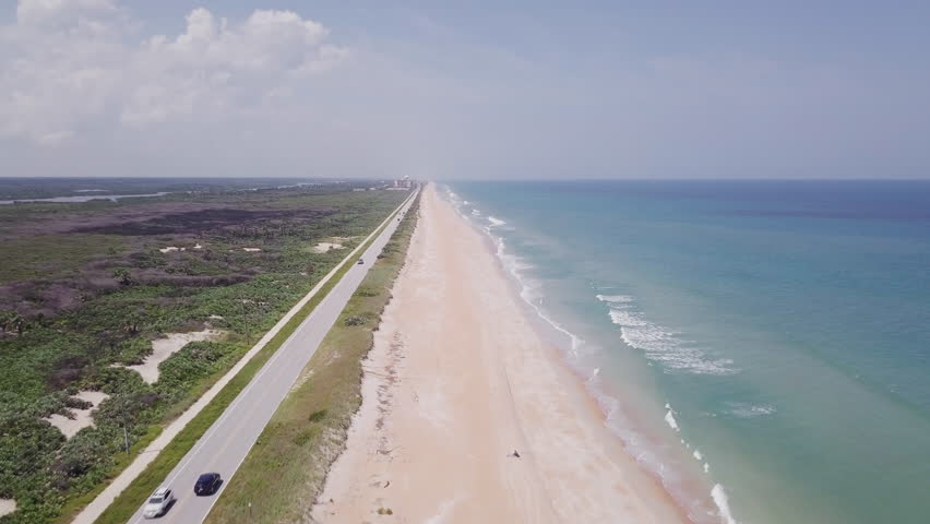 Aerial view on the Florida coastline. Road running parallel to the beach. Several cars rushing by. Waves crashing on the ?sandy beach.