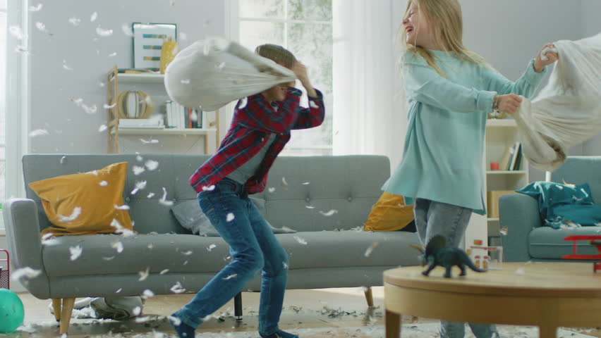 Adorable Little Boy and Sweet Little Girl Have a Pillow Fight in the Sunny Living Room. Siblings Having Fun Fighting with Pillows, Feathers Flying Around. In Slow Motion Royalty-Free Stock Footage #1021161139