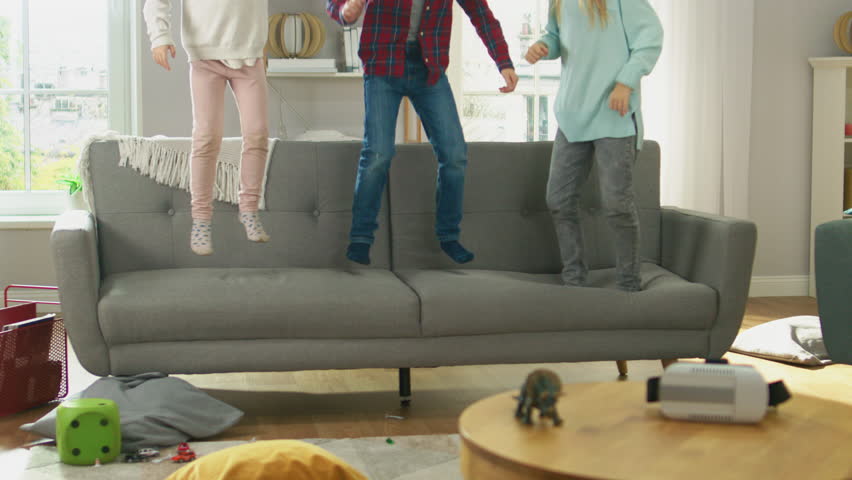 Two Cute Little Girls and Young Adorable Boy Have Fun, Jumping High on a Couch at Home. Happy Kids Dancing on a Sofa in the Sunny Living Room. In Slow Motion. Royalty-Free Stock Footage #1021161202