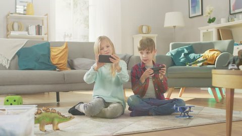 At Home Sitting on a Carpet: Cute Little Girl and Sweet Boy Playing Together in Video Game on two Smartphones, Holding them in Horizontal Landscape Mode. Happy Children do High Five.