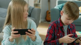 At Home Sitting on a Carpet: Cute Little Girl and Sweet Boy Playing in Competitive Video Game on two Smartphones, Holding them in Horizontal Landscape Mode. Close-up Portrait Camera Shot.