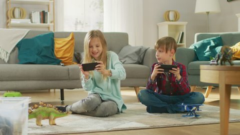 At Home Sitting on a Carpet: Cute Little Girl and Sweet Boy Playing in Competitive Video Game on two Smartphones, Holding them in Horizontal Landscape Mode. Girl Wins and Celebrates.