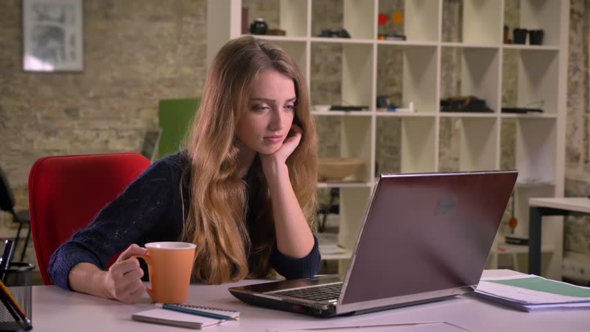 Portrait of pretty beautiful caucasian businesswoman delightfully watching on screen of laptop and drinking coffee in office. | Shutterstock HD Video #1021167943