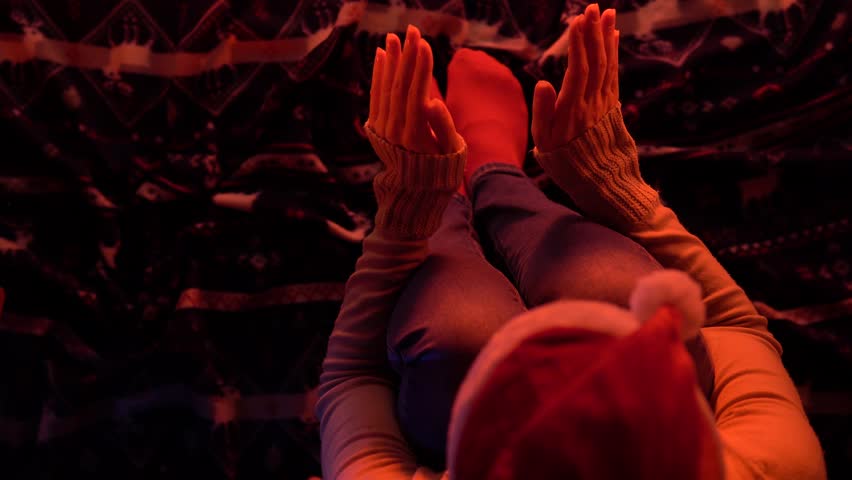 Anonymous woman in a Christmas hat sitting in front of the fireplace and basking, rubbing her hands against each other