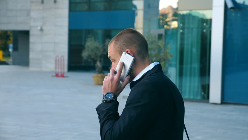 Businessman talking on phone near office and celebrating achievement. Young business man heard good news on cellphone and having positive emotions. Portrait of guy is happy with success. Dolly shot