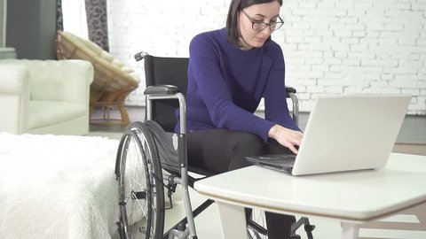young disabled woman with glasses in a wheelchair chair with a laptop at home,smiling