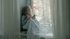 happy little girl with wavy red hair sits on the windowsill, covering a blanket and using the phone, talking, video calling 4k