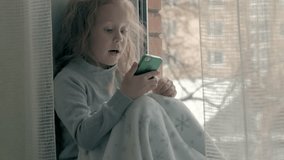 happy little girl with wavy red hair sitting on the windowsill, covering a blanket and using the phone, talking, video calling, close-up portrait 4k
