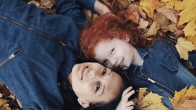 A beautiful mother and her cute daughter lie on a yellow leaf in the autumn park. They looking into the camera, top view. Yellow leaves falls on them. Happy family is having fun. Slow motion
