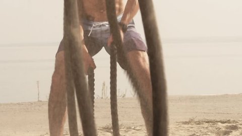 Slow Motion - Young Attractive Dynamic Handsome Man Battling Ropes Doing a Workout Exercise on a Beach. Bare Chested Man Exercising at the Seaside.