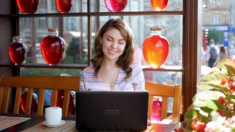 Smiling hipster girl talking and waving hand hello during online video call via laptop computer, sitting in coffee shop. Cheerful business woman having conference in internet via portable notebook