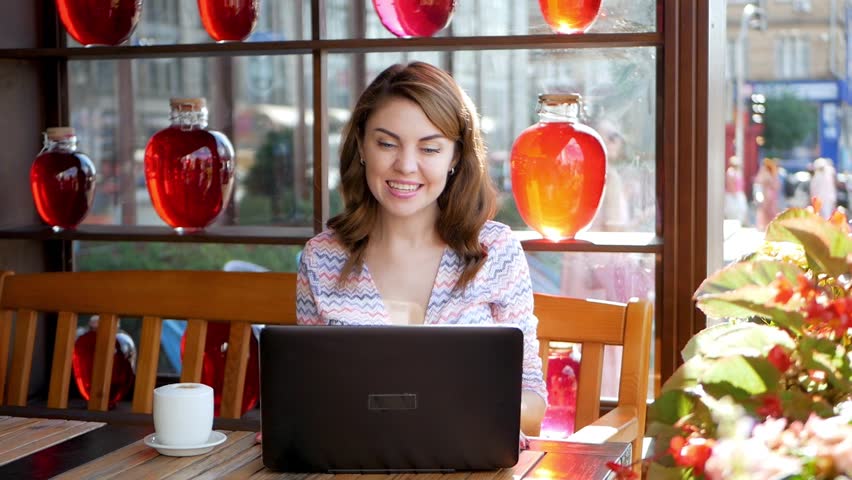 Smiling hipster girl talking and waving hand hello during online video call via laptop computer, sitting in coffee shop. Cheerful business woman having conference in internet via portable notebook Royalty-Free Stock Footage #1021211290
