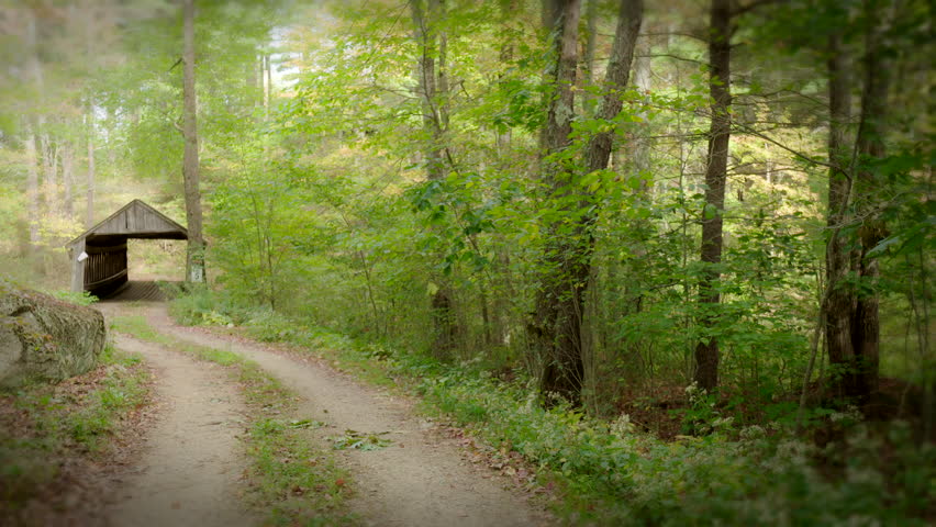 Covered bridge and dirt road in rural New England during autumn with fall foliage and leaves. Camera pans left.