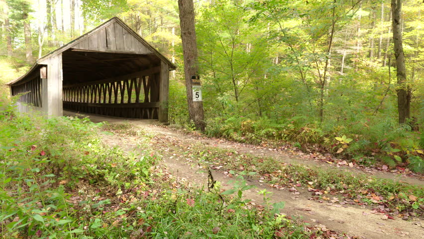 Old wooden covered bridge and dirt road in rural New England during autumn with fall foliage and leaves. Camera pans left. Soft focus with misty fog.