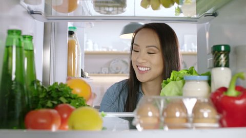 Young, happy Chinese woman opens a refrigerator and takes an apple for a healthy snack. POV from inside the refrigerator.