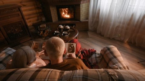 An elderly couple considers a family photo album wrapped in a blanket sitting by a cozy fireplace. Golden wedding, happy marriage, grow old together Video Stok