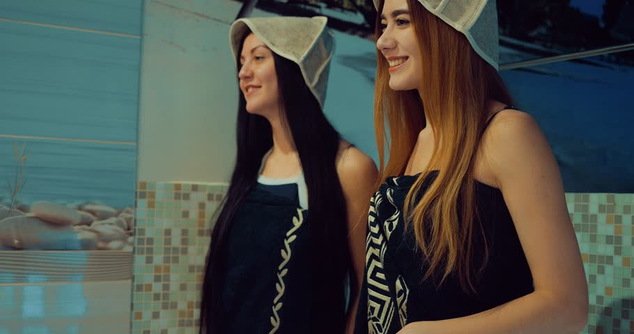Beautiful girlfriends in towels and bath hats come to the sauna. Open the glass door. Girls sit on a wooden bench. Beautiful girls are talking. Span camera.