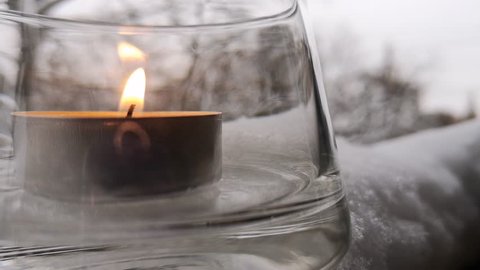 Closeup of a tealight candle in a transparent glass candlestick, which stands in the snow. Background in soft-focus.
