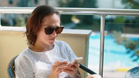 A woman in sunglasses with a smartphone sits at a glass table on the balcony. Woman reads SMS on the phone.