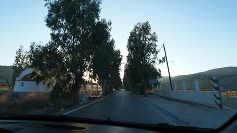 Drive at rural Cretan alley, entering small traditional village, evening sun light flash ahead. Car pass by small Church Agios Nektarios. Nice old trees grow along road