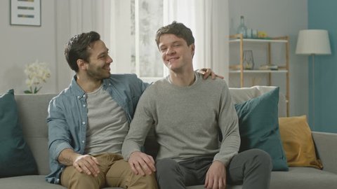 Cute Attractive Male Gay Couple Sit Together on a Sofa at Home. Boyfriend Puts His Hand on Partner's and They Hug. They are Happy and Smiling. They are Casually Dressed and Room Has Modern Interior.