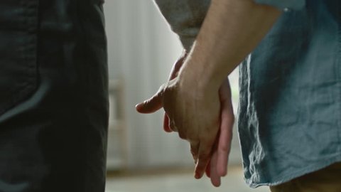 Close Up of Two Male Hands Gently Touching and Holding Each Other. Cute Queer Relationship Concept. Gay Couple are Casually Dressed. Room is Bright and Sunny.