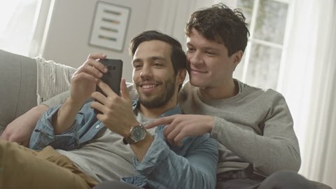 Cute Male Gay Couple Spend Time at Home. They are Lying Down on a Sofa and Use a Smartphone. They Browse Online. Partner's Hand is Around His Lover. They Smile and Laugh. Room Has Modern Interior.