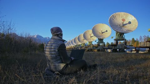 Hacker hacks the radio telescope using notebook. Man to program hacking on laptop in nature. Terrorist is hiding in camouflage balaclava. Hacker has criminal cases on the Internet. Programmer uses a