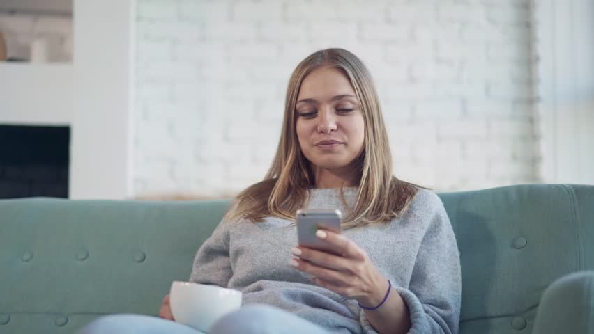 Attractive young woman with blond hair wearing gray is sitting on her sofa at home, drinking tea and web surfing on her smartphone. Handheld slow motion medium shot