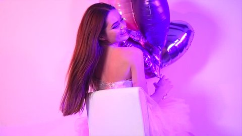 Beauty fashion model girl with heart shaped balloons having fun, sitting on chair, laughing and spinning. Emotions. Holiday celebration. Beautiful young brunette woman on party. Slow motion 4K UHD