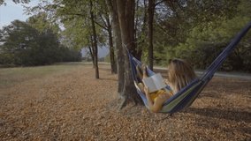 Cheerful blond girl on hammock reading book in nature enjoying peaceful environment- Female relaxing in summer 