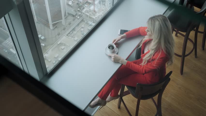 Woman in red coral business suit drinks coffee at a high table near the window. Young blonde businesswoman in the office in high floor in front of the window.
