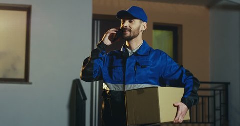 Caucasian handsome deliveryman in the blue uniform and cap standing at the house with carton box and talking on the phone. Outdoors.