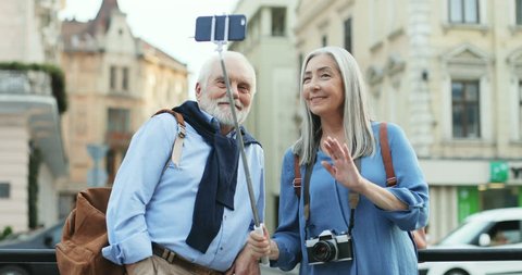 Caucasian old smiled couple of tourists standing in the city center with a smartphone on the selfie stick and having a videochat ot taking photos. Outside.