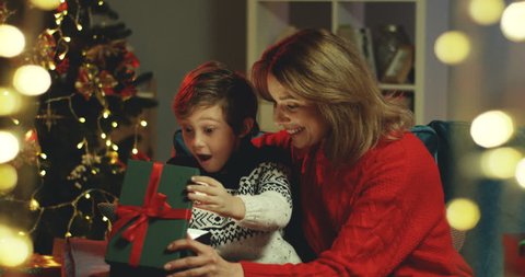 Caucasian beautiful mother and her son sitting at the Christmas tree and opening a present box with surprised and excited faces.
