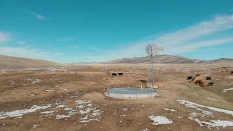 Aerial-Dolly right simulation past old fashioned windmill with cattle and light patches of snow on the desert hills on a clear winter day