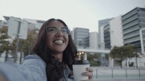 Smiling Latin woman taking selfie or making video call in city. Happy Hispanic tourist in glasses holding coffee to go. Selfie concept