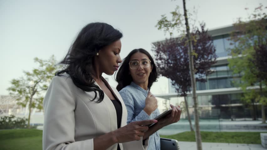 Confident coworkers with gadgets discussing work in park. Beautiful multicultural businesswomen with digital devices walking in city park. Remote work concept Royalty-Free Stock Footage #1021319779