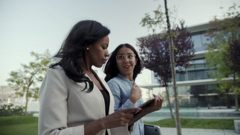 Confident coworkers with gadgets discussing work in park. Beautiful multicultural businesswomen with digital devices walking in city park. Remote work concept Stockvideo