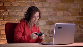 Portrait of young female blogger in red hoodie playing video game using joystick and commenting on bricken wall background.