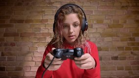 Close-up portrait of involved and interested blogger in headphones playing with joystick on bricken wall background.