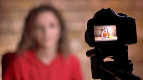 Close-up portrait of young blogger recording positive video in front of camera and lamp on bricken wall background.