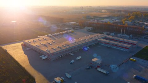 Aerial circling view of logistics center with warehouse, loading hub with many semi-trailers truck load/unload goods at sunset
