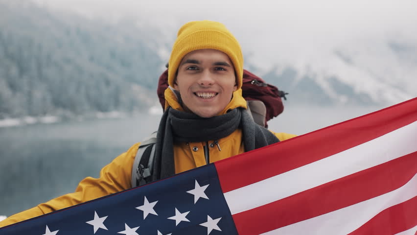 Traveller man holding the flag of America standing in the snow-covered mountains neat beautuful lake. He looking into the camera and smiling. Hiker traveller wearing in yellow winter clothes