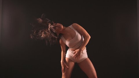 Beautiful hot brunette woman dancing sexy dance on black background in darkness. Young girl wears white short shorts, top and moves her hair