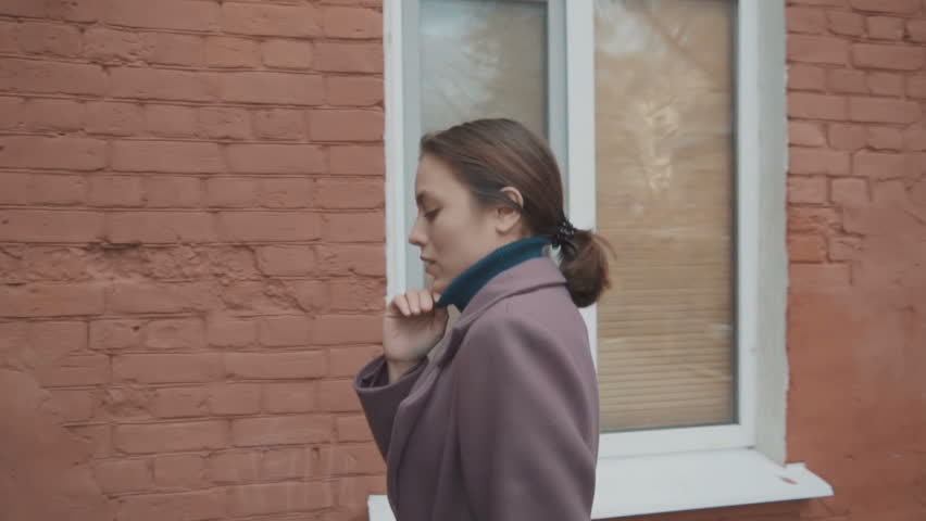 The girl in the lilac coat with a belt goes along the lanes between the houses and suddenly turns around and goes further. Slow motion.