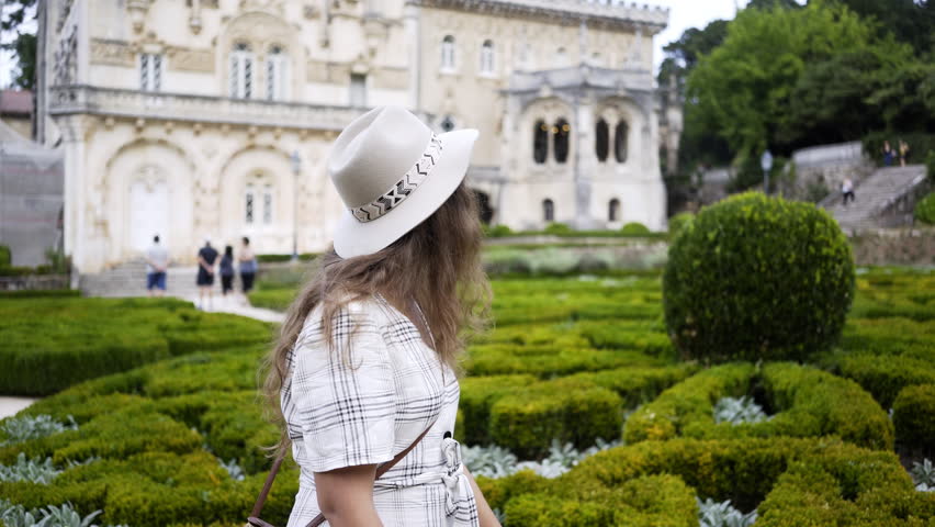 Side view of attractive young female in hat smiling and looking away while walking near green bushes in park of Bussaco Palace Hotel in Luso, Portugal