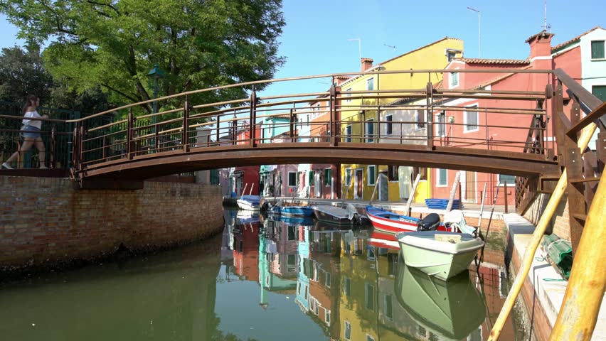 4K, Asian woman passing to a bridge of canal in street with colorful Venetian houses facade at island of Burano. Tourists visiting colourfully painted apartments buildings in the village-Dan