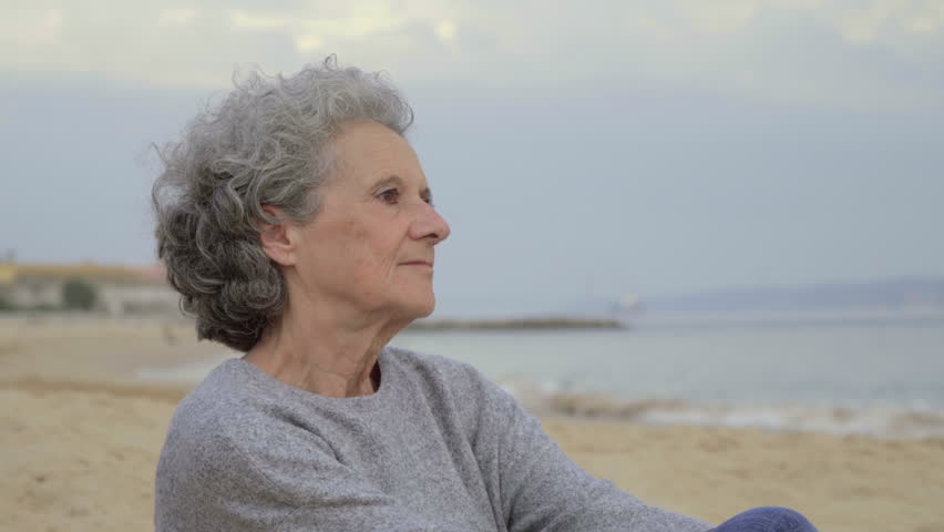 Thoughtful senior lady sitting on beach and looking at distance. Beautiful smiling woman wearing casual clothing looking at camera. Relaxation concept