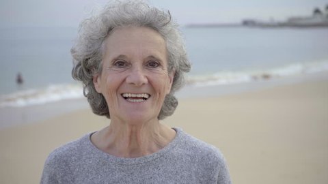 Happy beautiful elderly woman with toothy smile posing against blurred seascape background. Close up shot of smiling senior lady with brown eyes looking at camera. Happy retirement concept